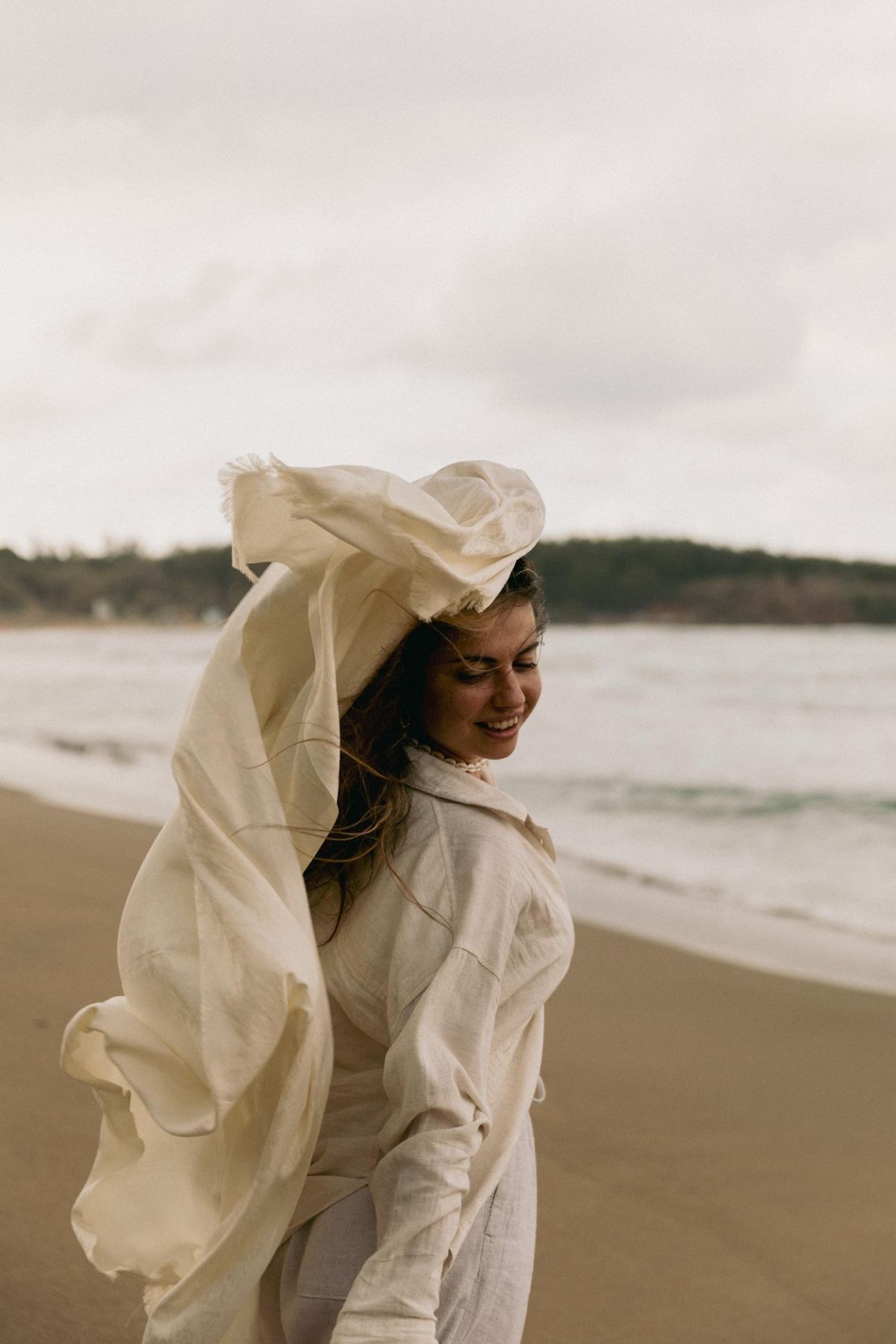 Lucia smiling on the beach as a cream scarf lifts over her head in the wind
