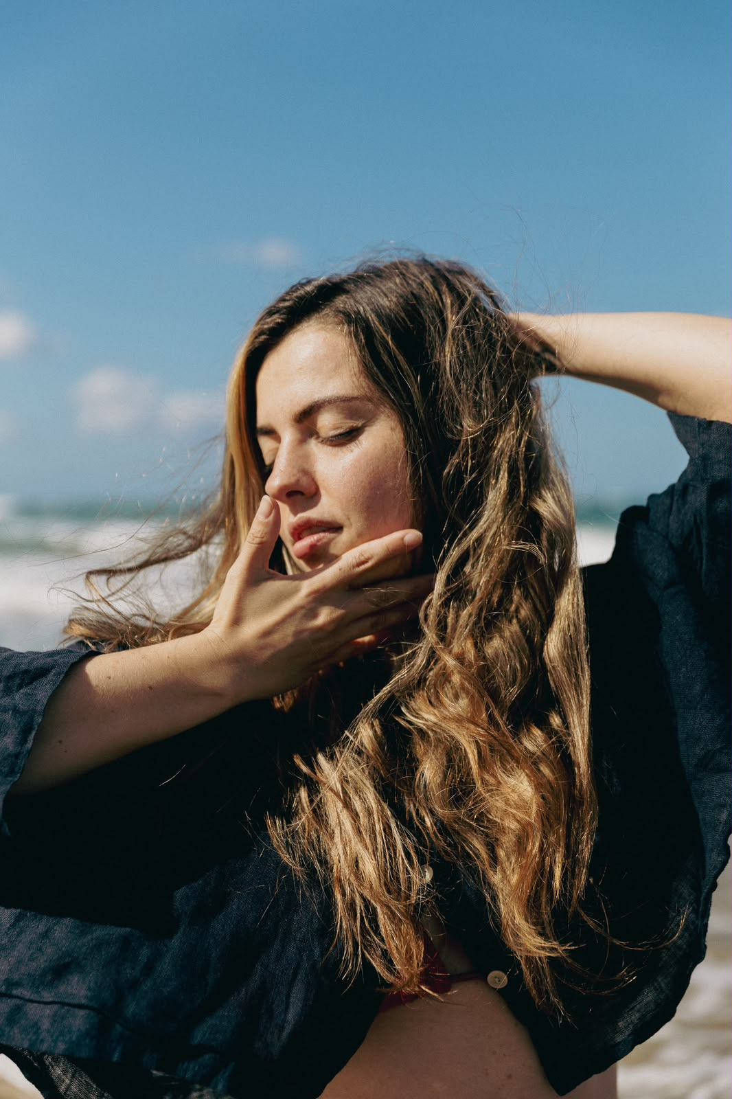 Lucia by the sea in the sun, eyes closed, one hand at her face and the other in her hair