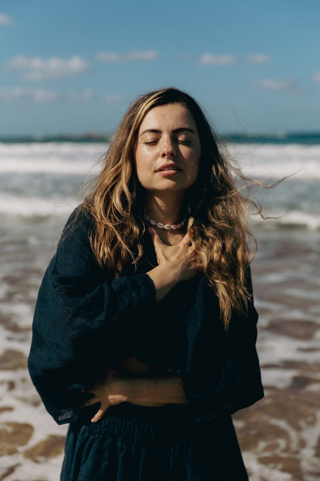 Lucia by the sea, eyes closed, one hand on her heart and one on her belly