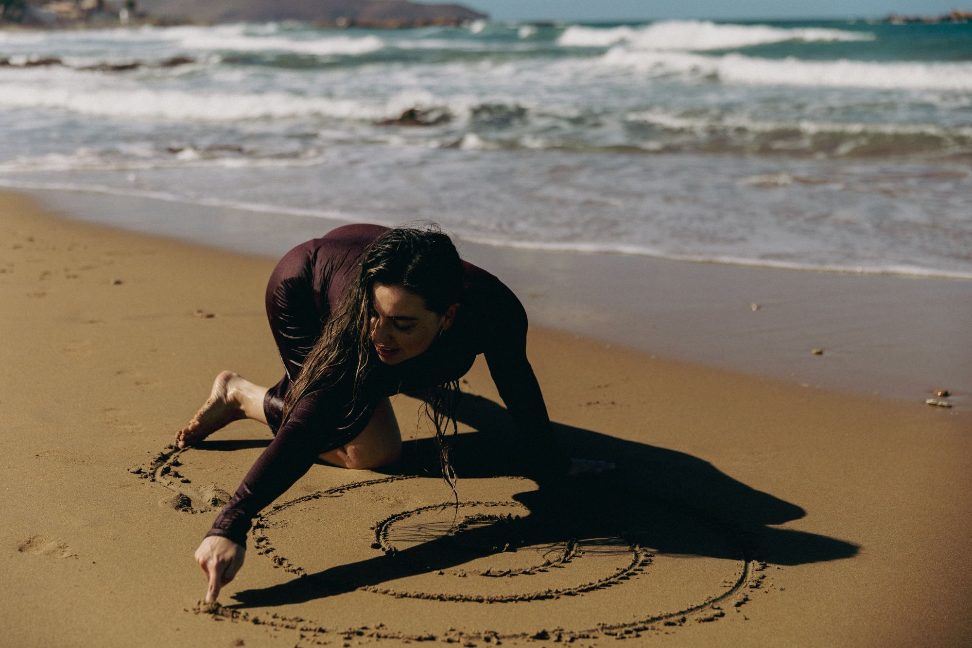 Lucia kneeling on a beach, drawing a spiral into the wet sand with her finger